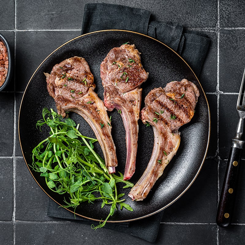 Grilled lamb chops with rocket leaves on a black plate.